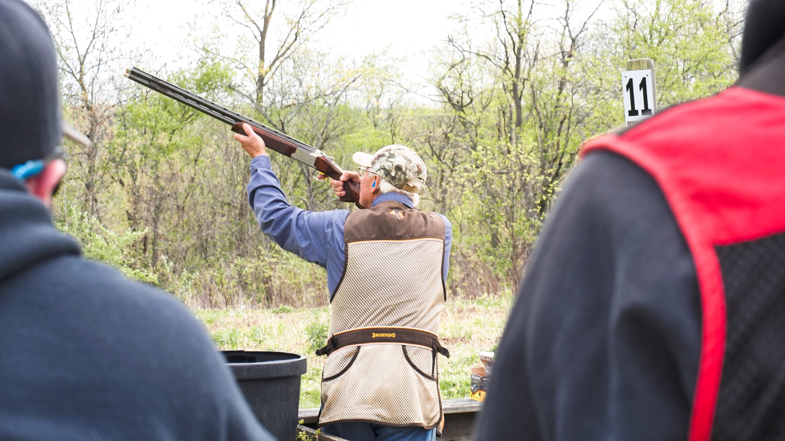 A person shoots a shotgun at a clay pigeon shooting range. The number 11 is visible on a post in the background.