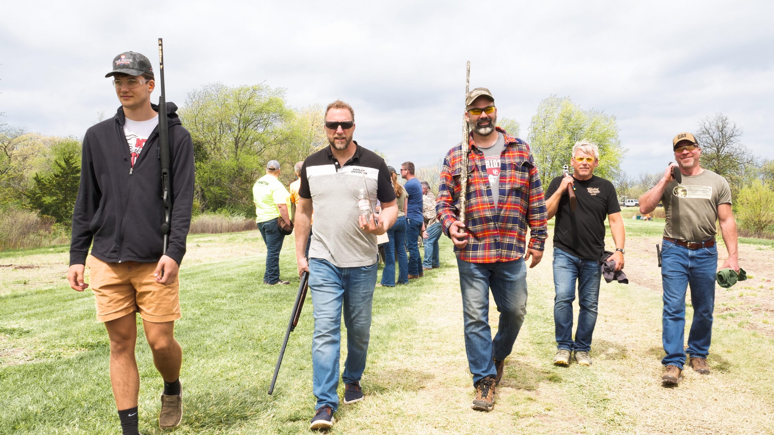 Five men walk across a grassy field, each carrying a shotgun.