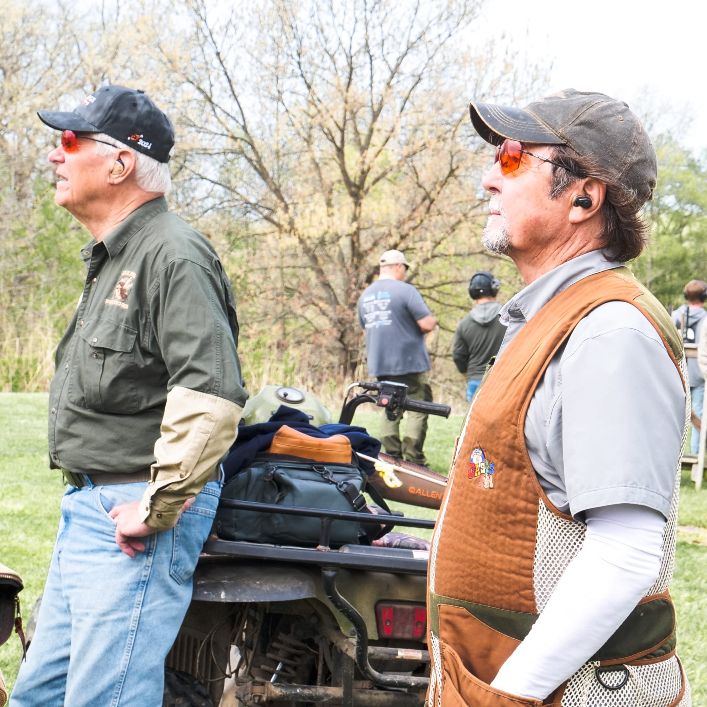 Two men wearing shooting vests and protective eyewear stand near an ATV at a shooting range.