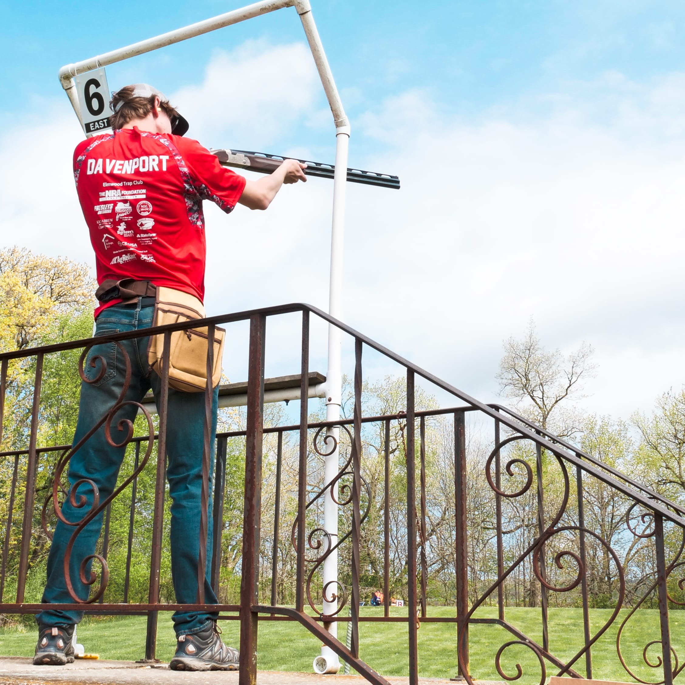 A person wearing a Davenport Elmwood Trap Club shirt shoots a shotgun at a trap range.